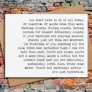 May include: A white card with black text on a brown envelope, set against a brick wall background. The card's message offers words of comfort and encouragement, including phrases like "Resting counts" and "You're not alone."