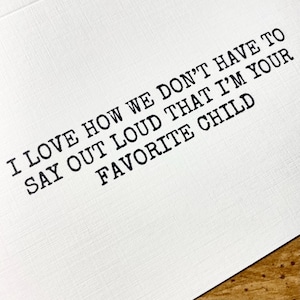 May include: White card with black text that reads: "I LOVE HOW WE DON'T HAVE TO SAY OUT LOUD THAT I'M YOUR FAVORITE CHILD." The text is in a typewriter-style font. The card is on a wooden surface.