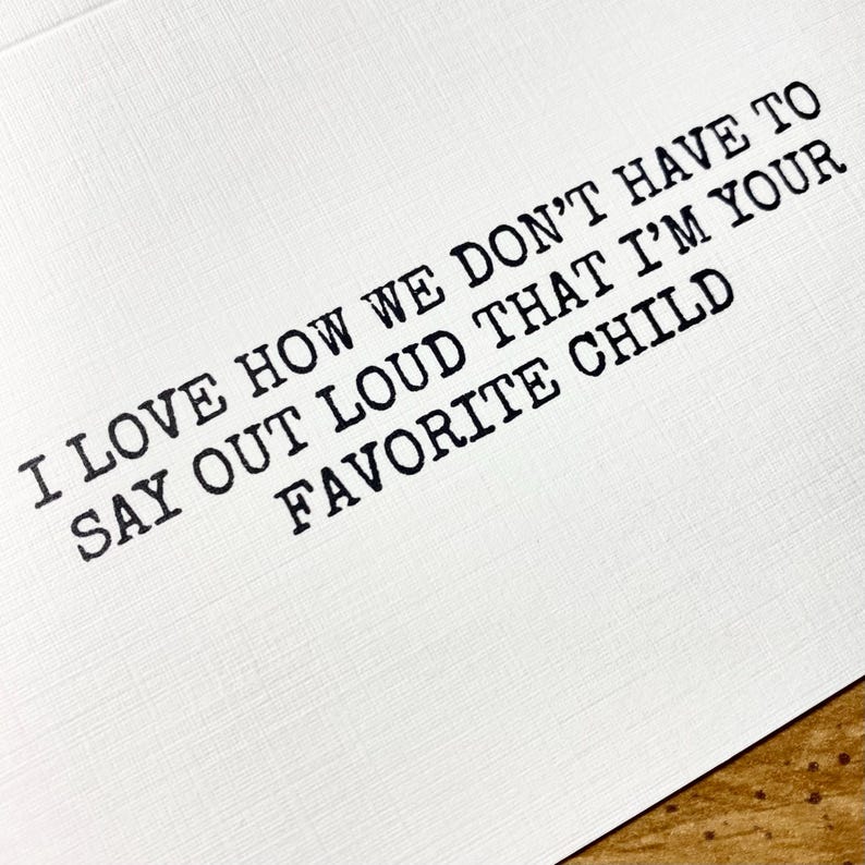 May include: White card with black text that reads: "I LOVE HOW WE DON'T HAVE TO SAY OUT LOUD THAT I'M YOUR FAVORITE CHILD." The text is in a typewriter-style font. The card is on a wooden surface.