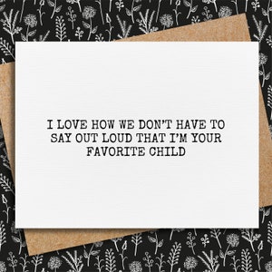 May include: A white greeting card with the text "I LOVE HOW WE DON'T HAVE TO SAY OUT LOUD THAT I'M YOUR FAVORITE CHILD" in black. The card is on a brown envelope, set against a black background with a white floral pattern.