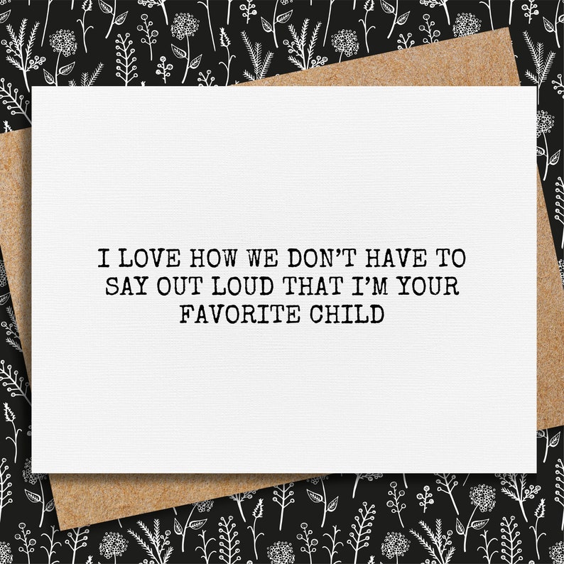 May include: A white greeting card with the text "I LOVE HOW WE DON'T HAVE TO SAY OUT LOUD THAT I'M YOUR FAVORITE CHILD" in black. The card is on a brown envelope, set against a black background with a white floral pattern.