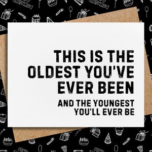 May include: A white greeting card with the text "THIS IS THE OLDEST YOU'VE EVER BEEN AND THE YOUNGEST YOU'LL EVER BE" in bold black letters. The card is inside a brown envelope, set against a black background with white birthday-themed illustrations.
