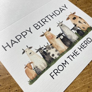 K&ouml;nnte beinhalten: Eine Gl&uuml;ckwunschkarte mit wei&szlig;em Hintergrund und schwarzem Text, der "Happy Birthday from the Herd" lautet. Die Karte zeigt eine Aquarell-Illustration einer Gruppe von K&uuml;hen und einem Schaf.