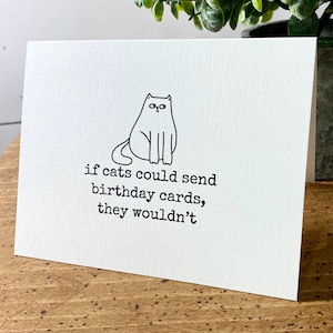 May include: A white card with a black line drawing of a cat sitting on it. The text on the card reads "if cats could send birthday cards, they wouldn't".