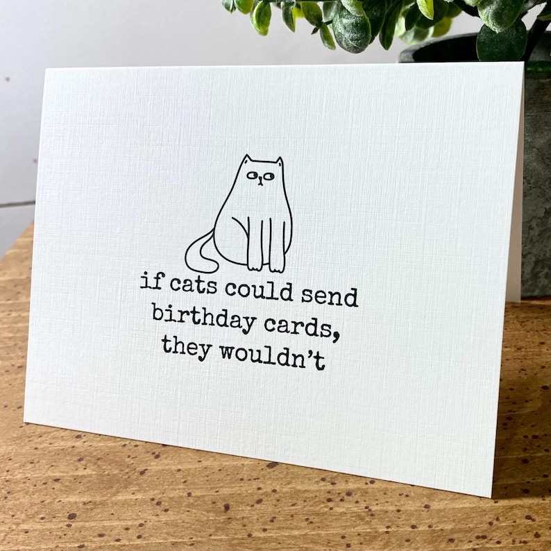 May include: A white card with a black line drawing of a cat sitting on it. The text on the card reads "if cats could send birthday cards, they wouldn't".