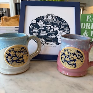 May include: Two ceramic mugs with a Hawaii theme. One is light blue and white, the other is pink and purple. Both mugs have a circular design with the word "Aloha" and a map of Hawaii. A framed print of the Hawaii map is in the background.