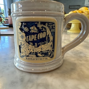 May include: A white ceramic mug with a blue and yellow design featuring a map of Cape Cod, Massachusetts. The mug has a handle and says "Cape Cod" and "Massachusetts" on the front.