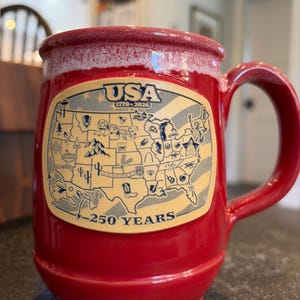 May include: A red ceramic mug with a speckled white rim featuring a tan rectangular graphic of the United States map. The design includes the text USA 1776-2026 and 250 YEARS, along with various small black icons representing landmarks and regional symbols.