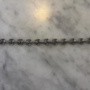 Puede incluir: Pulsera plateada con un diseño repetido de langosta. La pulsera está compuesta por múltiples eslabones en forma de langosta conectados entre sí, con un cierre de broche. La joya se muestra sobre una superficie de mármol.