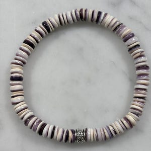 Puede incluir: Un brazalete de cuentas con cuentas de concha blancas y moradas alternas. El brazalete tiene una cuenta decorativa de color plateado. Las cuentas son redondas y de tamaño uniforme, creando una forma circular.