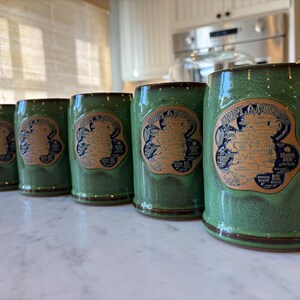 May include: A set of five green ceramic mugs with a brown rim and handle. Each mug features a tan label with the word "IRELAND" and a map design. The mugs are arranged on a white marble surface.