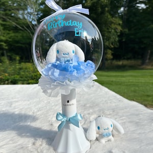 May include: A clear bubble with a white plush toy inside, surrounded by blue flowers. The bubble is on a white stand with a blue ribbon. The bubble has the text "Happy Birthday Erin" on it. There is a smaller white plush toy on the table.