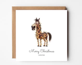 May include: A square greeting card featuring a brown horse wrapped in glowing yellow string lights. The text Merry Christmas and the name LOUISA are printed in black below the illustration. The card is paired with a brown kraft paper envelope.