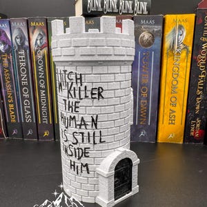 Puede incluir: Un sujetalibros gris en forma de castillo con el texto "Witch Killer The Human Is Still Inside Him". Una figura de lobo blanco descansa sobre un libro que dice "Blink, Blink, Blink, Blink". El sujetalibros est&aacute; frente a una fila de libros.
