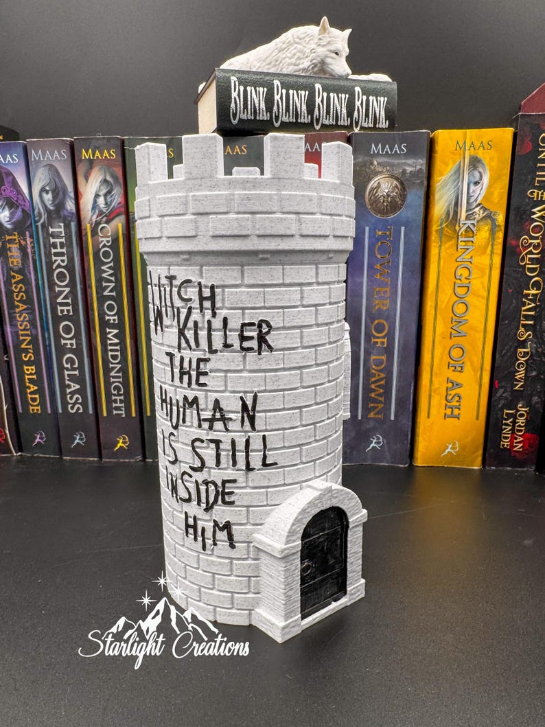 Puede incluir: Un sujetalibros gris en forma de castillo con el texto "Witch Killer The Human Is Still Inside Him". Una figura de lobo blanco descansa sobre un libro que dice "Blink, Blink, Blink, Blink". El sujetalibros est&aacute; frente a una fila de libros.