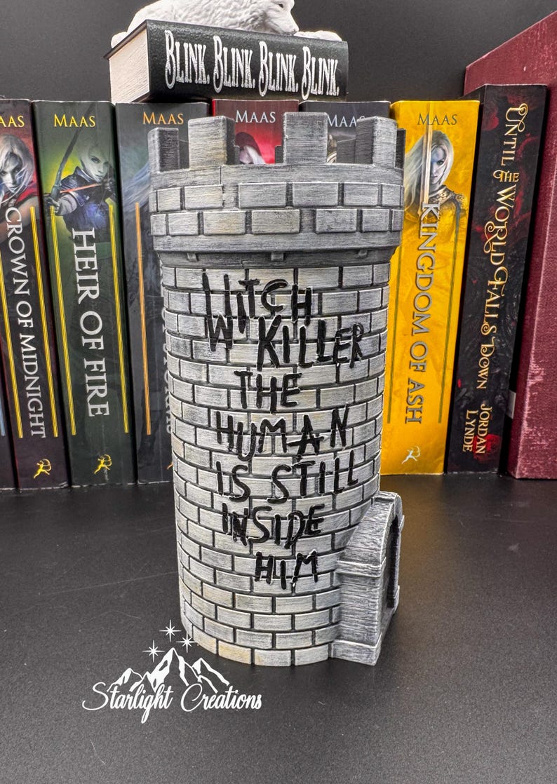 Puede incluir: Un sujetalibros gris en forma de castillo con el texto "Witch W Killer The Human Is Still Inside Him". El sujetalibros est&aacute; hecho de un material similar al ladrillo y tiene una peque&ntilde;a torre en el lateral. Libros en el fondo.
