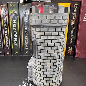 Puede incluir: Una figura de torre de castillo cil&iacute;ndrica gris con un patr&oacute;n de ladrillos, una peque&ntilde;a puerta arqueada y una parte superior almenada. La base tiene el texto "Starlight Creations". Libros con los t&iacute;tulos "Crown of Midnight", "Heir of Fire" y "Empire of Storms" est&aacute;n en el fondo.