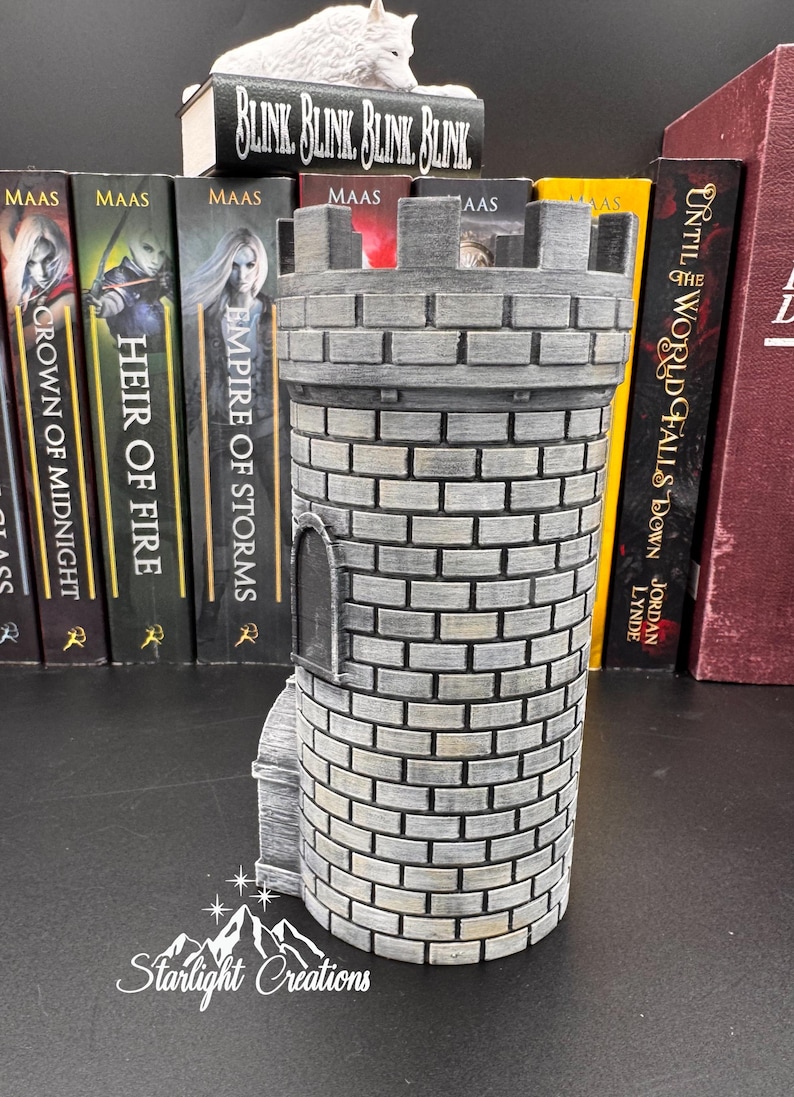 Puede incluir: Una figura de torre de castillo cil&iacute;ndrica gris con un patr&oacute;n de ladrillos, una peque&ntilde;a puerta arqueada y una parte superior almenada. La base tiene el texto "Starlight Creations". Libros con los t&iacute;tulos "Crown of Midnight", "Heir of Fire" y "Empire of Storms" est&aacute;n en el fondo.