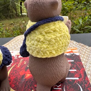 May include: A brown bear figurine wearing a yellow and navy blue crocheted shawl. The bear is holding a small, rectangular object. The figurine is sitting on top of a book with red and white text.