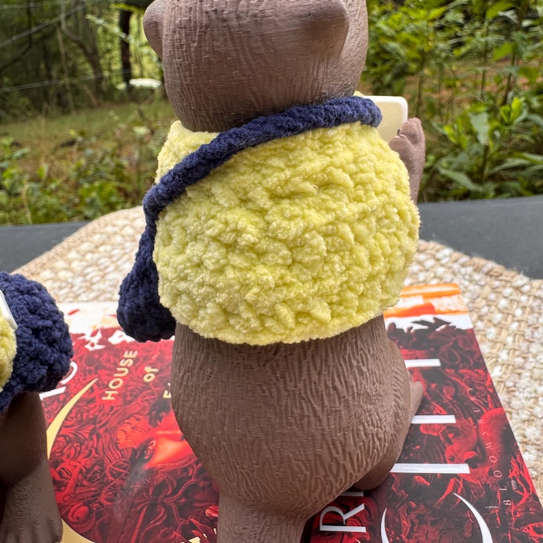 May include: A brown bear figurine wearing a yellow and navy blue crocheted shawl. The bear is holding a small, rectangular object. The figurine is sitting on top of a book with red and white text.