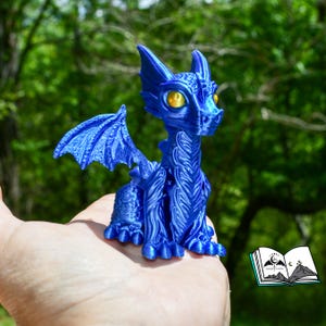 May include: A detailed, blue dragon figurine with textured wings and body. The dragon has yellow eyes and is posed in a seated position. The figurine is held in a hand against a blurred green background.