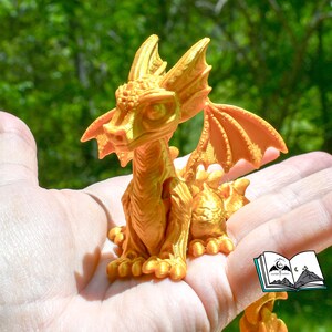 May include: A golden-colored dragon figurine with detailed wings, horns, and scales. The dragon is perched on a hand, showcasing its intricate design. The background is a blurred view of green foliage.