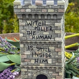 May include: A stone-textured tower sculpture with a crenelated top and a dark arched doorway. The exterior features hand-painted gray, tan, and brown brick patterns with the black text: WITCH KILLER THE HUMAN IS STILL INSIDE HIM.