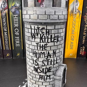Puede incluir: Un sujetalibros cil&iacute;ndrico gris en forma de castillo con el texto "Witch in Killer, The Human Is Still Inside Him". El sujetalibros est&aacute; hecho de un material similar al ladrillo y tiene una peque&ntilde;a puerta arqueada. Libros con la palabra "MAAS" est&aacute;n en el fondo.