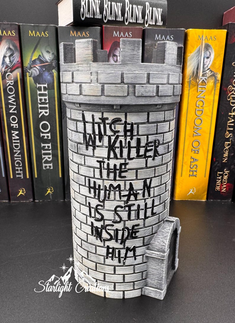Puede incluir: Un sujetalibros cil&iacute;ndrico gris en forma de castillo con el texto "Witch in Killer, The Human Is Still Inside Him". El sujetalibros est&aacute; hecho de un material similar al ladrillo y tiene una peque&ntilde;a puerta arqueada. Libros con la palabra "MAAS" est&aacute;n en el fondo.