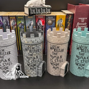 Puede incluir: Cuatro recipientes decorativos en forma de torre en tonos grises y verde azulado, cada uno con una parte superior almenada y una peque&ntilde;a puerta arqueada. El texto "Witch Killer The Human Is Still Inside Him" est&aacute; escrito en cada torre. Libros y una peque&ntilde;a escultura en el fondo.