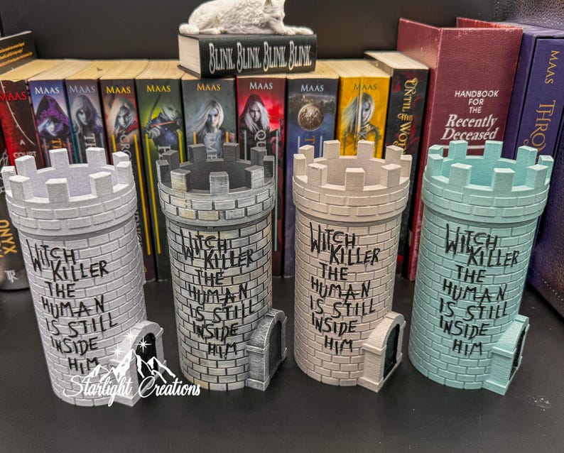 Puede incluir: Cuatro recipientes decorativos en forma de torre en tonos grises y verde azulado, cada uno con una parte superior almenada y una peque&ntilde;a puerta arqueada. El texto "Witch Killer The Human Is Still Inside Him" est&aacute; escrito en cada torre. Libros y una peque&ntilde;a escultura en el fondo.