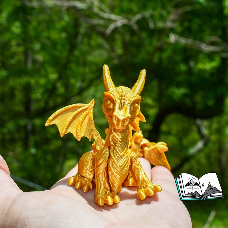 May include: A golden dragon figurine with detailed wings, horns, and scales. The dragon is sitting with its front paws extended. The figurine is held in a hand, with a blurred green background.