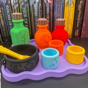 May include: A set of colorful potion bottles, small cups, and a black cauldron with a yellow spoon, all arranged on a wavy purple tray. The items feature embossed symbols including a leaf, moon, heart, water drop, and flower, with brown cork stoppers.