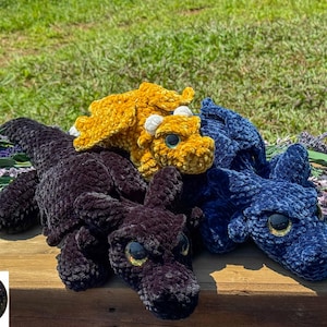 May include: Three plush crochet dragon toys in black, mustard yellow, and dark blue. Each dragon features large, glossy, reflective eyes, textured yarn bodies, and small wings. A circular logo with the text Starborn Hollow is in the corner.