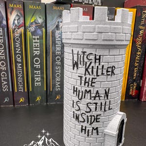 Puede incluir: Una torre de castillo gris con el texto "Witch Killer The Human Is Still Inside Him" en negro. La torre tiene una peque&ntilde;a puerta y est&aacute; hecha de un material similar al ladrillo. Hay libros en el fondo.