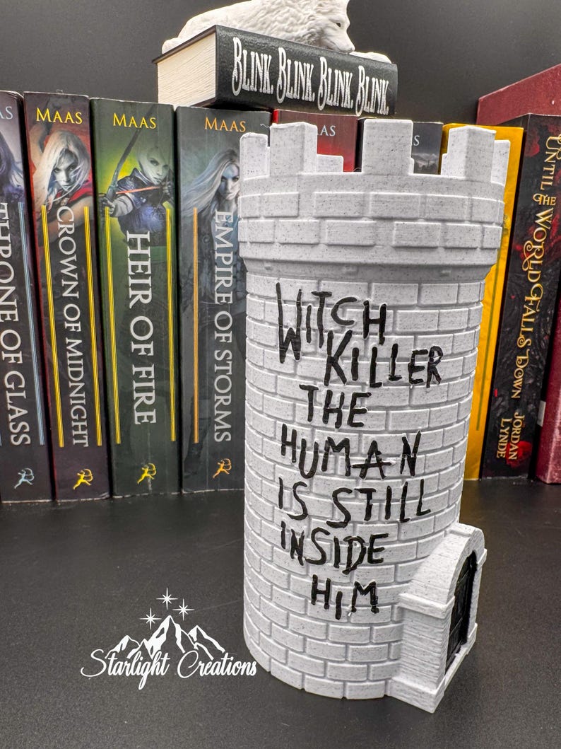 Puede incluir: Una torre de castillo gris con el texto "Witch Killer The Human Is Still Inside Him" en negro. La torre tiene una peque&ntilde;a puerta y est&aacute; hecha de un material similar al ladrillo. Hay libros en el fondo.