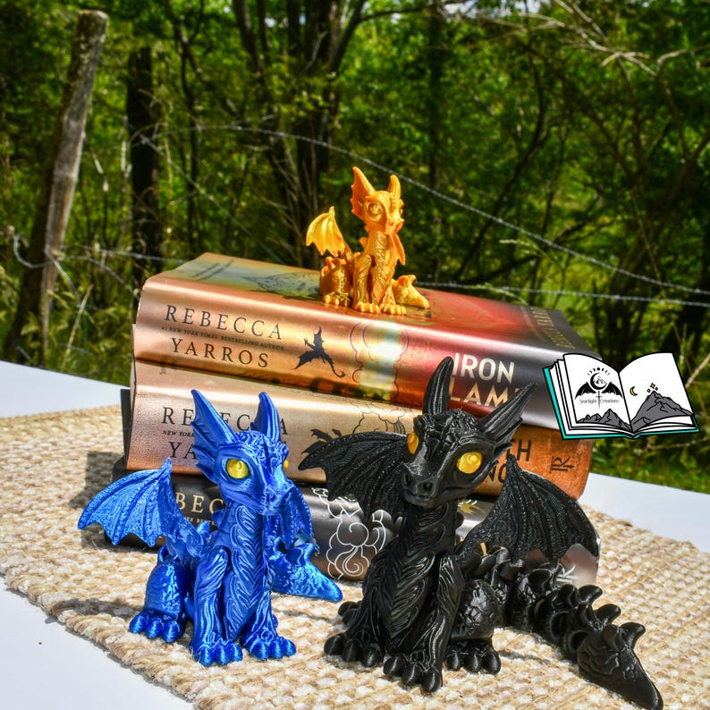 May include: A collection of fantasy-themed figurines, including a blue dragon, a black dragon, and a gold dragon. The dragons are posed in front of a stack of books with the author's name "Rebecca Yarros" visible. A small illustration of a book is in the background.