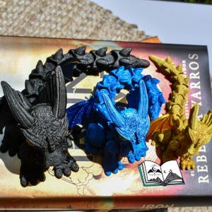 May include: Three detailed dragon figurines in black, blue, and gold rest on a book. The dragons have textured scales, wings, and horns. The book has the title "Rebel" and the author's name. A small logo of a book with a moon and mountains is visible.