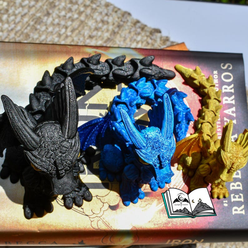 May include: Three detailed dragon figurines in black, blue, and gold rest on a book. The dragons have textured scales, wings, and horns. The book has the title "Rebel" and the author's name. A small logo of a book with a moon and mountains is visible.