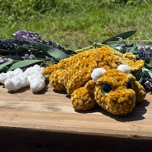 May include: A golden-yellow crocheted plush dragon toy with a soft, velvet-like texture. The creature features large, dark, reflective eyes and small white horns on its head. Beside the dragon are several small, white, fluffy crocheted cloud shapes.