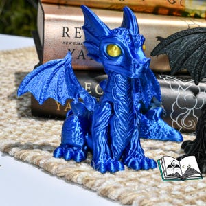 May include: A vibrant blue dragon figurine with detailed wings and textured scales. The dragon has yellow eyes and is posed sitting upright. The figurine is on a neutral-toned surface, with books in the background.