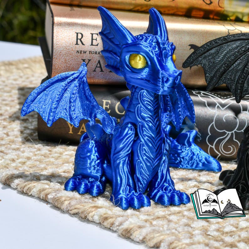 May include: A vibrant blue dragon figurine with detailed wings and textured scales. The dragon has yellow eyes and is posed sitting upright. The figurine is on a neutral-toned surface, with books in the background.