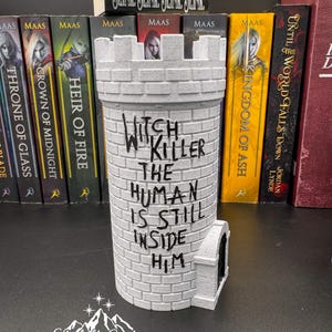 Puede incluir: Un objeto decorativo gris en forma de castillo con el texto "Witch Killer The Human Is Still Inside Him". El art&iacute;culo est&aacute; hecho de un material similar al ladrillo y tiene una peque&ntilde;a puerta arqueada. Libros y una figura de lobo en el fondo.