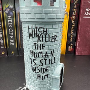 Puede incluir: Un objeto decorativo en forma de castillo azul claro con el texto "Witch, Will Killer, The Human Is Still Inside Him". El objeto tiene una peque&ntilde;a puerta y una textura similar a ladrillos. Hay libros en el fondo.