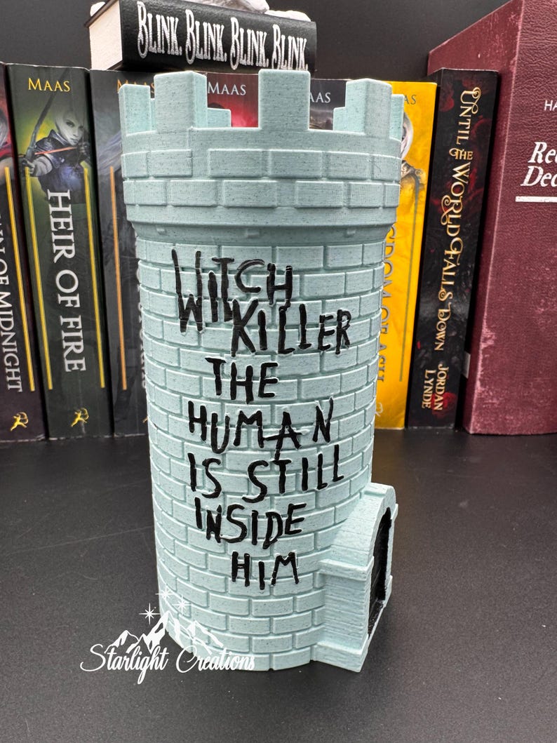 Puede incluir: Un objeto decorativo en forma de castillo azul claro con el texto "Witch, Will Killer, The Human Is Still Inside Him". El objeto tiene una peque&ntilde;a puerta y una textura similar a ladrillos. Hay libros en el fondo.