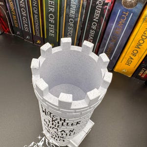 Puede incluir: Un portal&aacute;pices en forma de castillo gris claro con el texto "Witch Killer Human Still Inside Him". El portal&aacute;pices tiene una parte superior almenada y una base rectangular. El logotipo de Starlight Creations es visible en la parte inferior. Libros en el fondo.