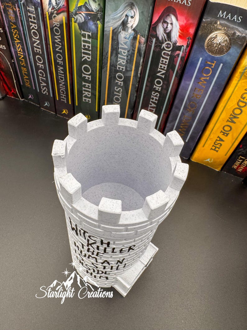 Puede incluir: Un portal&aacute;pices en forma de castillo gris claro con el texto "Witch Killer Human Still Inside Him". El portal&aacute;pices tiene una parte superior almenada y una base rectangular. El logotipo de Starlight Creations es visible en la parte inferior. Libros en el fondo.