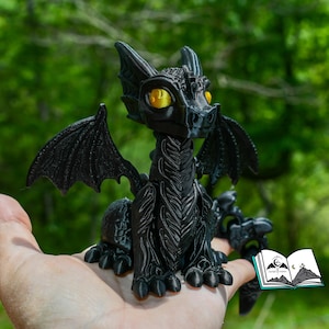 May include: A black dragon figurine with yellow eyes and detailed wings. The dragon is sitting, with its wings spread. The figurine is held in a hand, with a blurred green background. The dragon is approximately 4 inches tall.