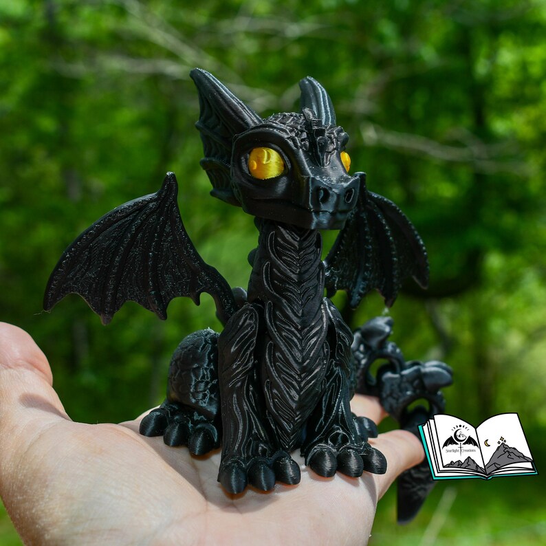 May include: A black dragon figurine with yellow eyes and detailed wings. The dragon is sitting, with its wings spread. The figurine is held in a hand, with a blurred green background. The dragon is approximately 4 inches tall.