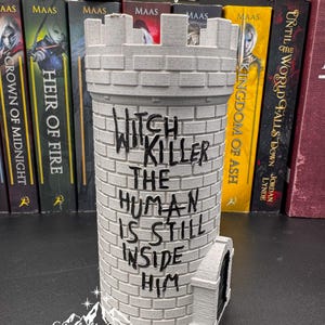 Puede incluir: Un objeto decorativo gris en forma de castillo con el texto "Witch Killer The Human Is Still Inside Him". El objeto tiene una parte superior almenada y una peque&ntilde;a puerta arqueada. Hay libros en el fondo.